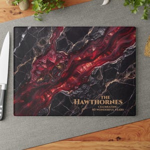 May include: A rectangular glass cutting board featuring a black marble pattern with a prominent, jagged red crystal vein running diagonally across the centre. The text reads THE HAWTHORNES CELEBRATING 40 WONDERFUL YEARS in gold lettering.