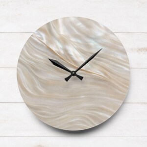 May include: A round wall clock featuring a face with a shimmering, iridescent mother-of-pearl finish displaying flowing, wavy patterns in shades of cream and off-white. The clock has two sharp, black metal hands centred on the dial.