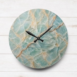 May include: A circular wall clock featuring a light blue and white marbled stone pattern with gold-toned veining throughout the face. The clock has two black pointed hands centred on the dial, indicating the time without any numerical markers.