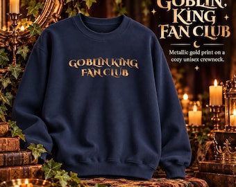 May include: A navy blue crewneck sweatshirt featuring the text "GOBLIN KING FAN CLUB" printed in a metallic gold font across the chest. The garment has long sleeves, a ribbed neckline, and a standard fit, showcasing a clean and simple design.