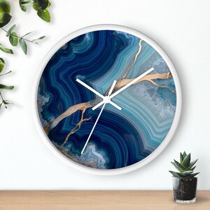 May include: A round wall clock with a white frame and a face featuring a blue and teal agate stone pattern with gold-coloured veining. The clock has three white hands positioned at the centre, displaying a circular design of layered mineral bands.