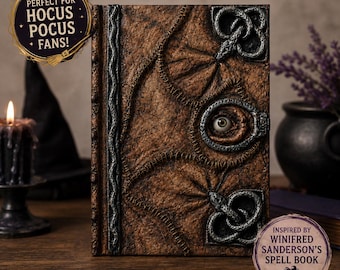 May include: A brown, textured book cover featuring a central human-like eye, silver-toned metal accents, and stitched detailing. The cover includes text reading "PERFECT FOR HOCUS POCUS FANS!" and "INSPIRED BY WINIFRED SANDERSON'S SPELL BOOK".
