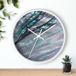 Op de afbeelding: Een ronde wandklok met een witte rand en witte wijzers. De wijzerplaat heeft een levendig, iriserend abalone schelppatroon met wervelende texturen in tinten van diep turkoois, glinsterend blauw, zacht paars en parelmoerachtige zilveren tinten.