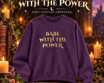 May include: A deep purple crewneck sweatshirt featuring the gold-colored text "BABE WITH THE POWER" printed on the center of the chest. The garment has long sleeves, a ribbed crew neckline, and ribbed cuffs, providing a standard fit and solid color.