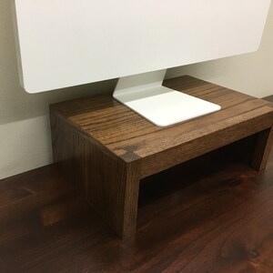 May include: A wooden monitor stand with a dark brown stain. The stand is made of solid wood and has a simple design. It is perfect for raising a computer monitor to a more ergonomic height.