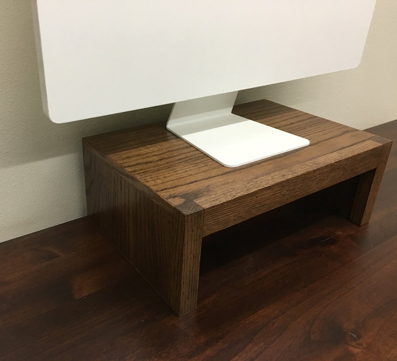 May include: A wooden monitor stand with a dark brown stain. The stand is made of solid wood and has a simple design. It is perfect for raising a computer monitor to a more ergonomic height.
