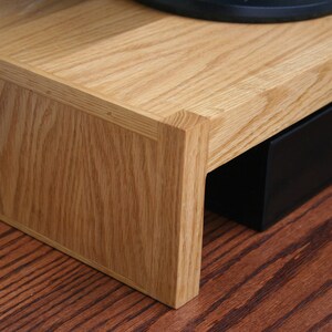 May include: A wooden stand with a drawer, made of light brown oak wood. The stand has a rectangular shape and a smooth surface. The drawer is black and is partially open.