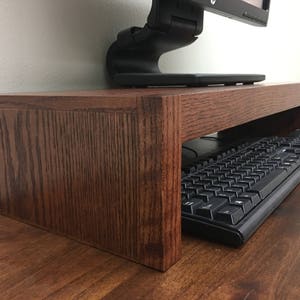 May include: A dark brown wooden monitor stand with a keyboard and mouse underneath. The stand is made of solid wood and has a simple design.