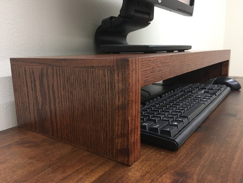 May include: A dark brown wooden monitor stand with a keyboard and mouse underneath. The stand is made of solid wood and has a simple design.