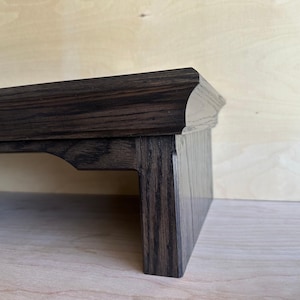 May include: A dark brown wooden monitor stand with a smooth, polished finish and a visible wood grain pattern. The stand features a flat top surface with a slightly curved edge and a sturdy vertical support leg with a decorative cutout detail.