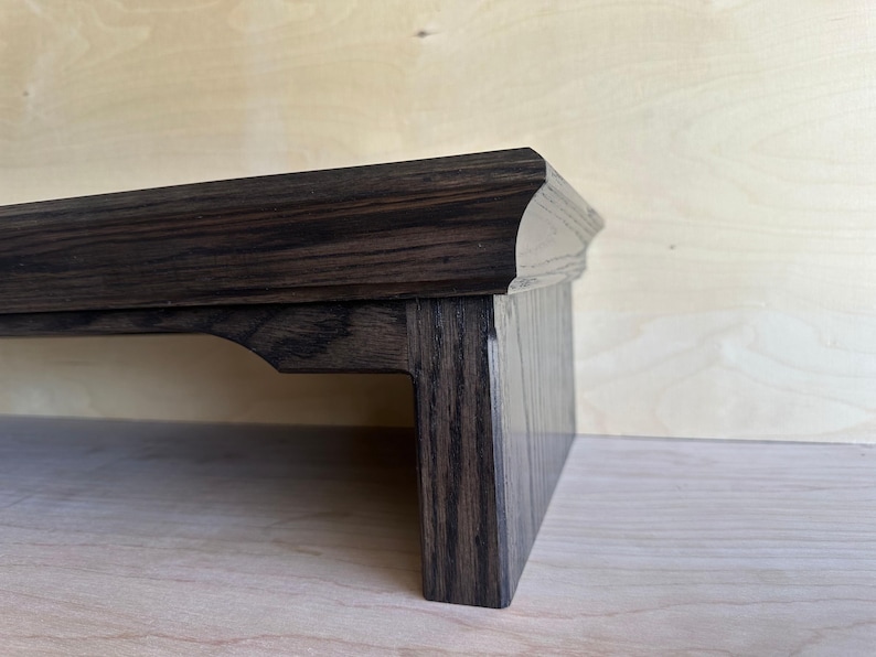 May include: A dark brown wooden monitor stand with a smooth, polished finish and a visible wood grain pattern. The stand features a flat top surface with a slightly curved edge and a sturdy vertical support leg with a decorative cutout detail.