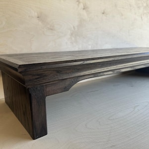 May include: A dark brown wooden monitor stand with a rectangular top surface and a visible wood grain texture. The stand features a raised edge detail along the perimeter and is supported by two sturdy legs with arched cutouts on the sides.