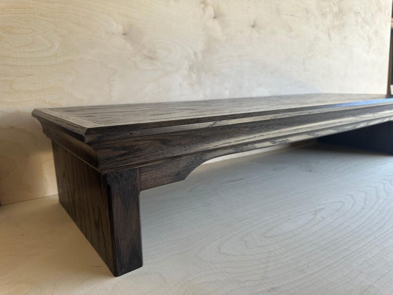 May include: A dark brown wooden monitor stand with a rectangular top surface and a visible wood grain texture. The stand features a raised edge detail along the perimeter and is supported by two sturdy legs with arched cutouts on the sides.
