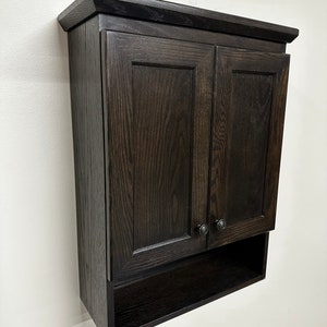 May include: A dark brown wooden wall cabinet with two doors and a shelf. The cabinet has a simple design with a slightly raised edge around the top and bottom.