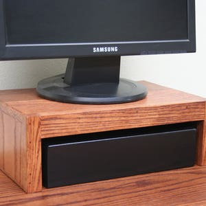 May include: A wooden monitor stand with a drawer, featuring a dark brown wood finish. The stand is holding a black computer monitor with the Samsung logo visible.
