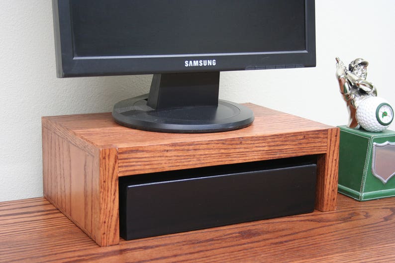 May include: A wooden monitor stand with a drawer, featuring a dark brown wood finish. The stand is holding a black computer monitor with the Samsung logo visible.