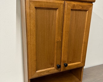 May include: A wall-mounted wooden cabinet featuring two shaker-style doors with black round knobs and a crown molding top. Below the doors is an open storage shelf. The cabinet has a medium-toned wood grain finish throughout.