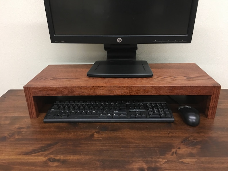 May include: A dark wood monitor stand with a black computer monitor, keyboard, and mouse on top. The stand has a shelf underneath for storage.