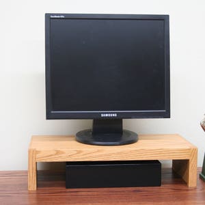 May include: A wooden monitor stand with a black box underneath. A black computer monitor sits on top of the stand. A small green and silver trophy is on the right side of the stand.