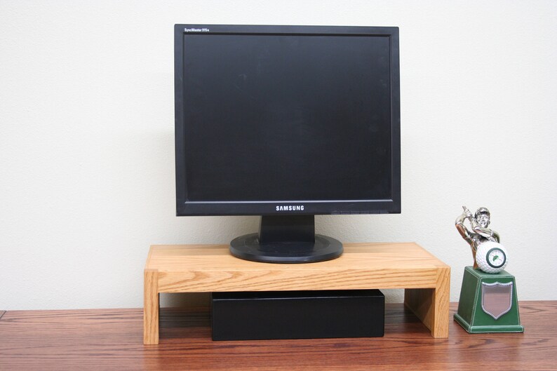 May include: A wooden monitor stand with a black box underneath. A black computer monitor sits on top of the stand. A small green and silver trophy is on the right side of the stand.