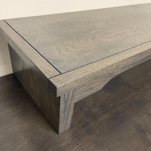 May include: A low-profile rectangular wooden stand featuring a dark gray stained finish with a visible wood grain pattern. The stand has a flat top surface, thick side panels, and a cutout detail on the lower edge of the support legs.