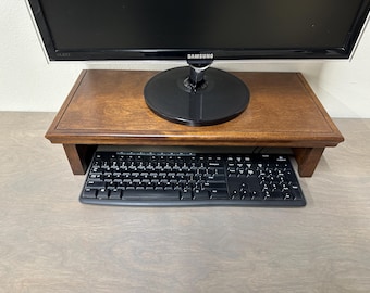 May include: A dark wood monitor stand with a keyboard underneath. The stand is raised off the surface and has a lip on the front to prevent items from falling off.