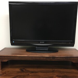 May include: A dark brown wooden TV stand with a flat top surface and a raised lip around the edge. The stand has a simple design with four legs and a rectangular shape. A black flat screen television is sitting on top of the stand.