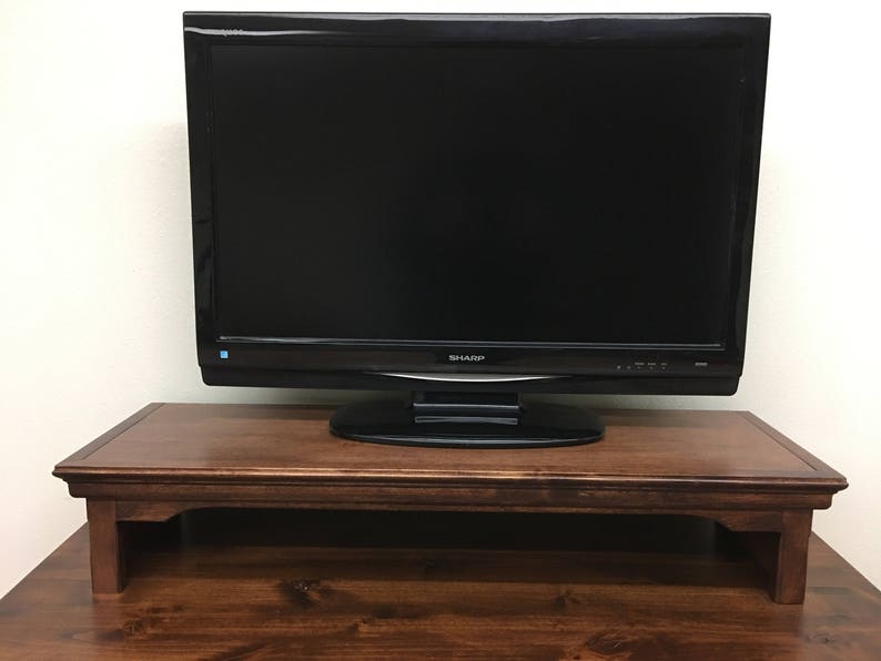 May include: A dark brown wooden TV stand with a flat top surface and a raised lip around the edge. The stand has a simple design with four legs and a rectangular shape. A black flat screen television is sitting on top of the stand.