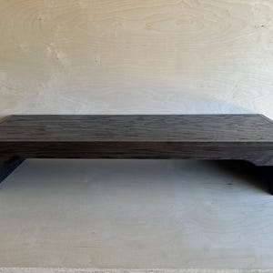 May include: A rectangular dark brown wooden display riser with a textured top surface and a beveled edge. The stand features four short, sturdy legs that provide elevation, creating a low-profile platform with a visible wood grain pattern.