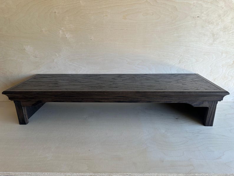May include: A rectangular dark brown wooden display riser with a textured top surface and a beveled edge. The stand features four short, sturdy legs that provide elevation, creating a low-profile platform with a visible wood grain pattern.