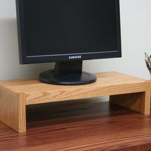 May include: A wooden monitor stand with a natural oak finish. The stand is raised off the surface and has a rectangular shape. A black computer monitor is sitting on top of the stand.