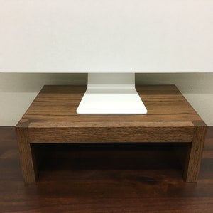 May include: A wooden monitor stand with a dark brown finish. The stand is made of solid wood and has a simple, modern design. It is designed to elevate a computer monitor to a more ergonomic height.
