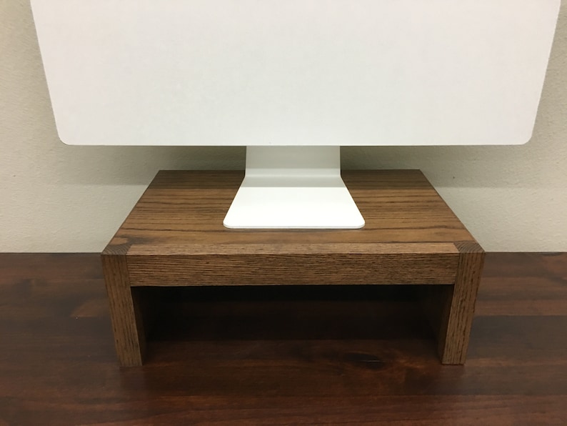 May include: A wooden monitor stand with a dark brown finish. The stand is made of solid wood and has a simple, modern design. It is designed to elevate a computer monitor to a more ergonomic height.