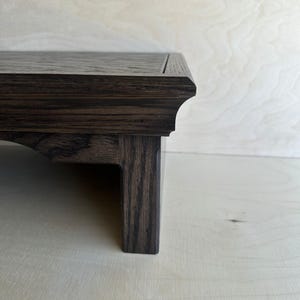 May include: A dark brown wooden display stand featuring a flat rectangular top with a beveled edge and a sturdy leg. The wood grain is visible throughout the surface, showcasing a deep, rich tone with a smooth finish and a clean, structured design.
