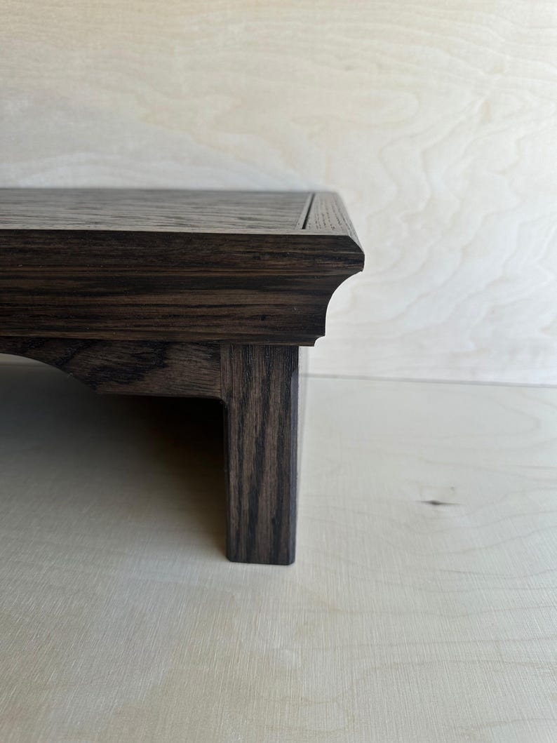 May include: A dark brown wooden display stand featuring a flat rectangular top with a beveled edge and a sturdy leg. The wood grain is visible throughout the surface, showcasing a deep, rich tone with a smooth finish and a clean, structured design.