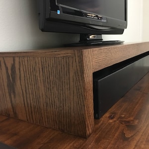 May include: A rectangular brown oak wood monitor stand with a visible wood grain pattern. The stand features an open space underneath, currently housing a black soundbar, and supports a black television screen on its top surface.