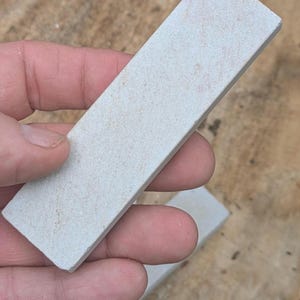 Puede incluir: Una piedra de afilar rectangular de color gris claro con una superficie ligeramente texturizada. La piedra se sostiene en una mano, mostrando su forma y tamaño. Herramienta para afilar cuchillos y otras hojas.