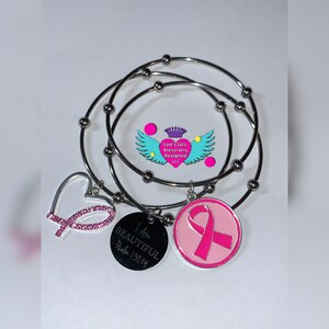 May include: Three silver bangle bracelets with charms. The charms include a pink ribbon, a heart with pink glitter, and a round charm with the words "I am beautiful - Psalm 139:14".