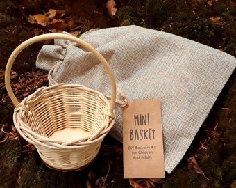 May include: A small, natural woven wicker basket with a wooden handle. The basket is sitting on a brown, mossy surface with a brown paper tag that reads "MINI BASKET DIY Basketry Kit for Children And Adults".