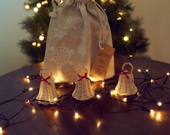 May include: Three small woven wicker bells with red ribbon bows, sitting on a wooden table with a string of white Christmas lights. A burlap bag with a snowflake pattern and a tag that reads "Christmas Bells, Natural" is behind the bells.