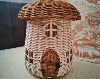 May include: A whimsical, woven wicker mushroom house in shades of light brown and cream. The house features a domed roof, a round window, and a small door with a black knob. The design is reminiscent of a fairy tale cottage.