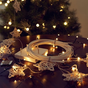 May include: A festive scene with Christmas lights and star-shaped ornaments. A wooden table displays a bundle of lights, star-shaped decorations, and a wooden crafting tool. A Christmas tree with glowing lights is in the background.