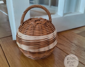 May include: A small, round woven basket with a handle. The basket is primarily brown with a wide band of natural beige around the middle. The basket sits on a wooden surface. The basket has a woven handle.
