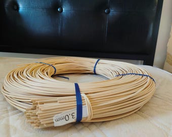 May include: A coiled bundle of light beige round reed material for weaving, secured with blue plastic bands. A white label attached to the front of the coil clearly displays the text 3.0mm, indicating the diameter of the individual reed strands.