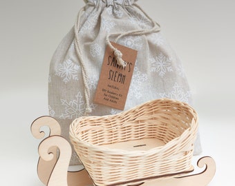 May include: A wooden sleigh with a wicker basket on top. The sleigh is natural wood and the basket is woven. A white fabric bag with snowflakes printed on it is behind the sleigh. The bag has a tag that says "Santa's Sleigh Natural DIY Basketry Kit for Children and Adults".