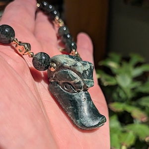 May include: A dark green stone necklace with a duck pendant. The pendant is intricately carved, and the necklace includes gold-coloured accents and dark green beads. The necklace is held in a hand.