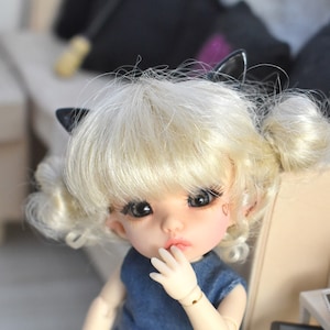 Può includere: Una piccola bambola con i capelli biondi raccolti in due codini, indossa un vestito intero di jeans e orecchie di gatto nere. La bambola si tiene una mano alla bocca e guarda la fotocamera.