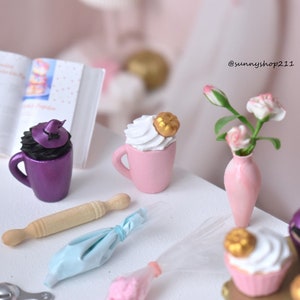 Può includere: Una scena da cucina in miniatura con una tazza viola con un cappello da strega nero, una tazza rosa con panna montata bianca e una zucca dorata, un vaso rosa con fiori rosa, un mattarello di legno, una sac à poche blu e due cupcake con cappelli da strega neri.
