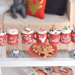 Può includere: Un set di otto tazze rosse in miniatura con scritte bianche e vari disegni a tema invernale, tra cui "happy fall y'all", "let it snow", "stay cozy", "winter", "hello winter", "warm winter wishes" e una renna. Le tazze sono disposte su un tavolo di vetro con un piatto di biscotti di pan di zenzero.