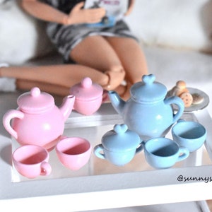 Könnte beinhalten: Ein rosa-blaues Miniatur-Tee-Set mit Teekanne, Zuckerdose und Tassen. Das Tee-Set steht auf einem weißen Tisch mit Glasplatte. Das Tee-Set ist perfekt für ein Puppenhaus oder ein Spielset.
